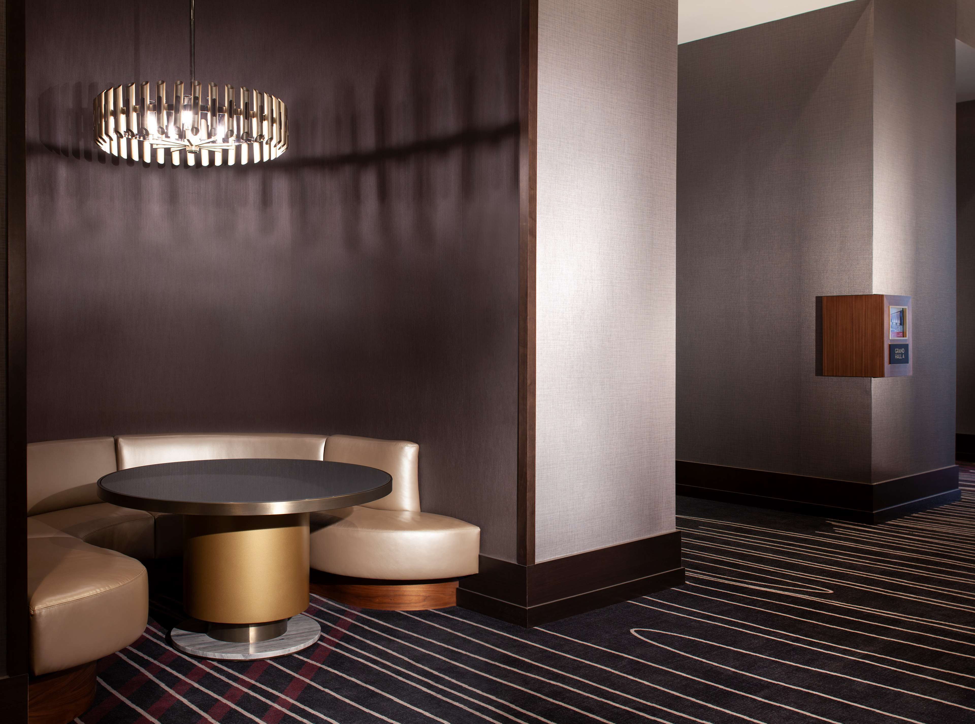 A modern hotel lobby with a round table, leather seating, a unique chandelier, and a wall-mounted information panel.