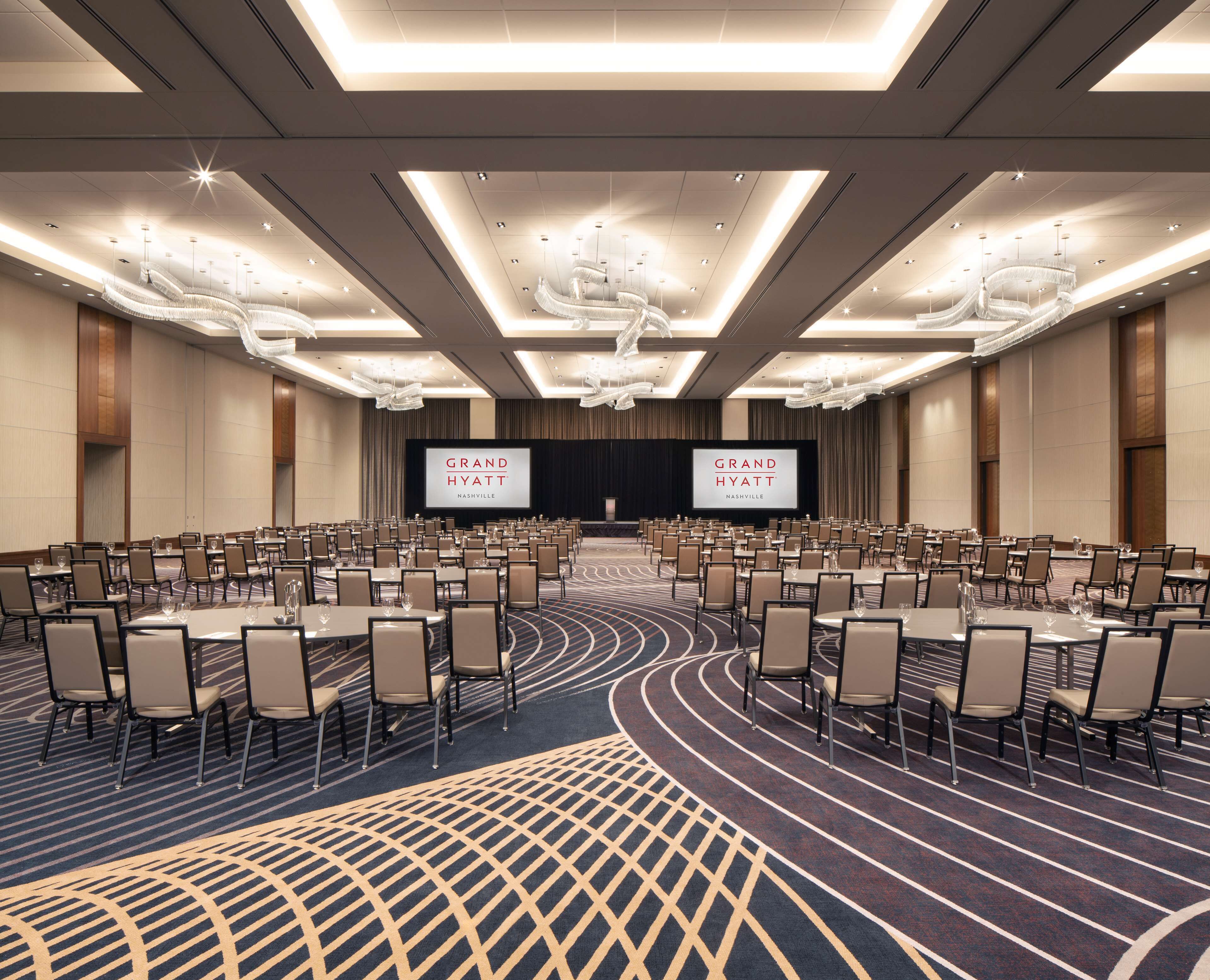 Un amplio salón de conferencias con filas de sillas, un escenario con una pantalla que muestra 'Grand Hyatt Nashville' y modernas lámparas de araña.