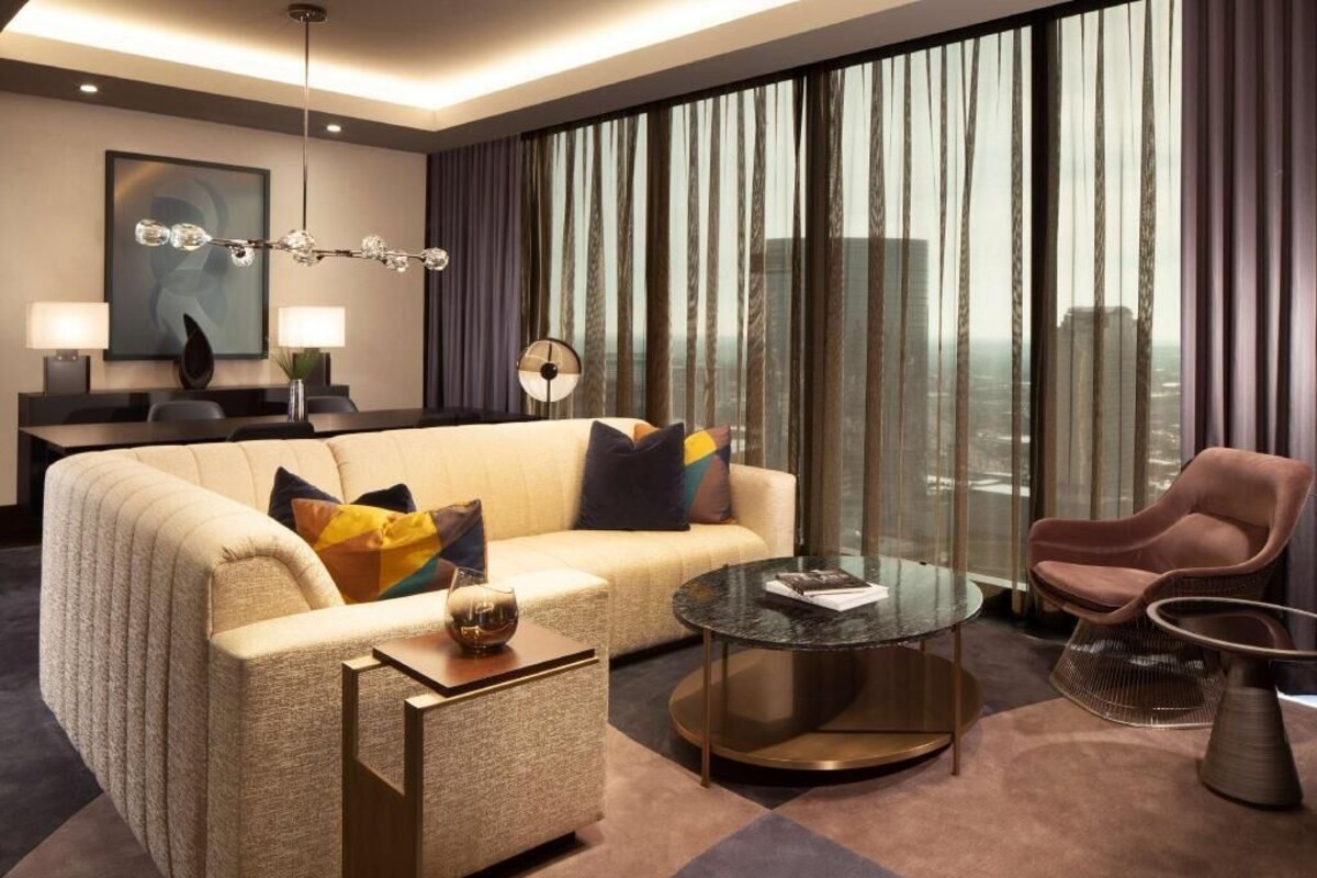 Una habitación de hotel moderna con sofá, una mesita redonda, una silla, escritorio con lámpara y un gran ventanal con cortinas.