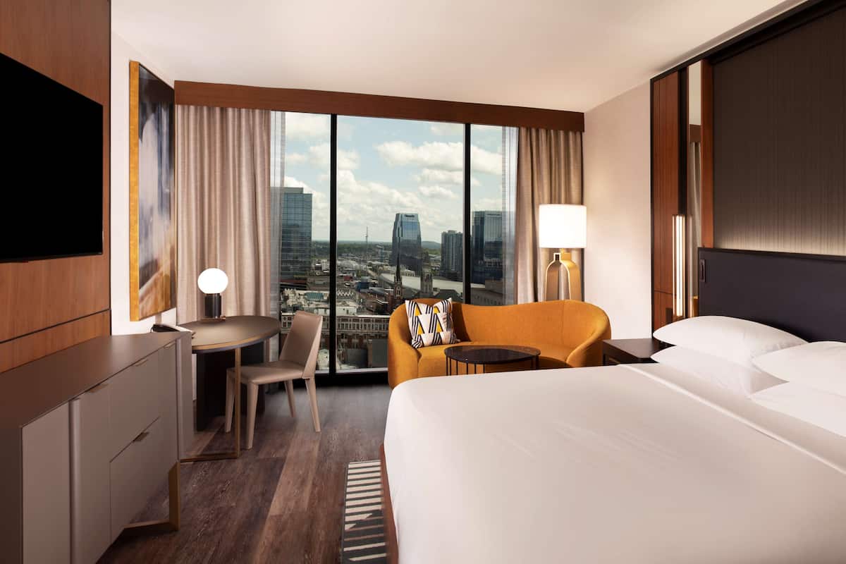 Een hotelkamer met een groot bed, een bureau, een stoel, een kleine tafel en een bank. De kamer heeft een groot raam met uitzicht op de stad.