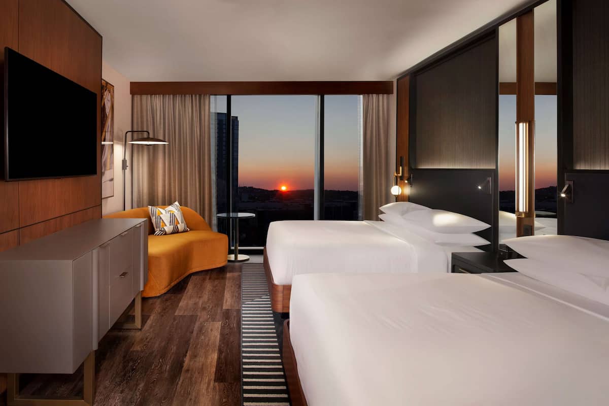 Een hotelkamer met een grote televisie, twee bedden, een sofa en uitzicht op de zonsondergang.