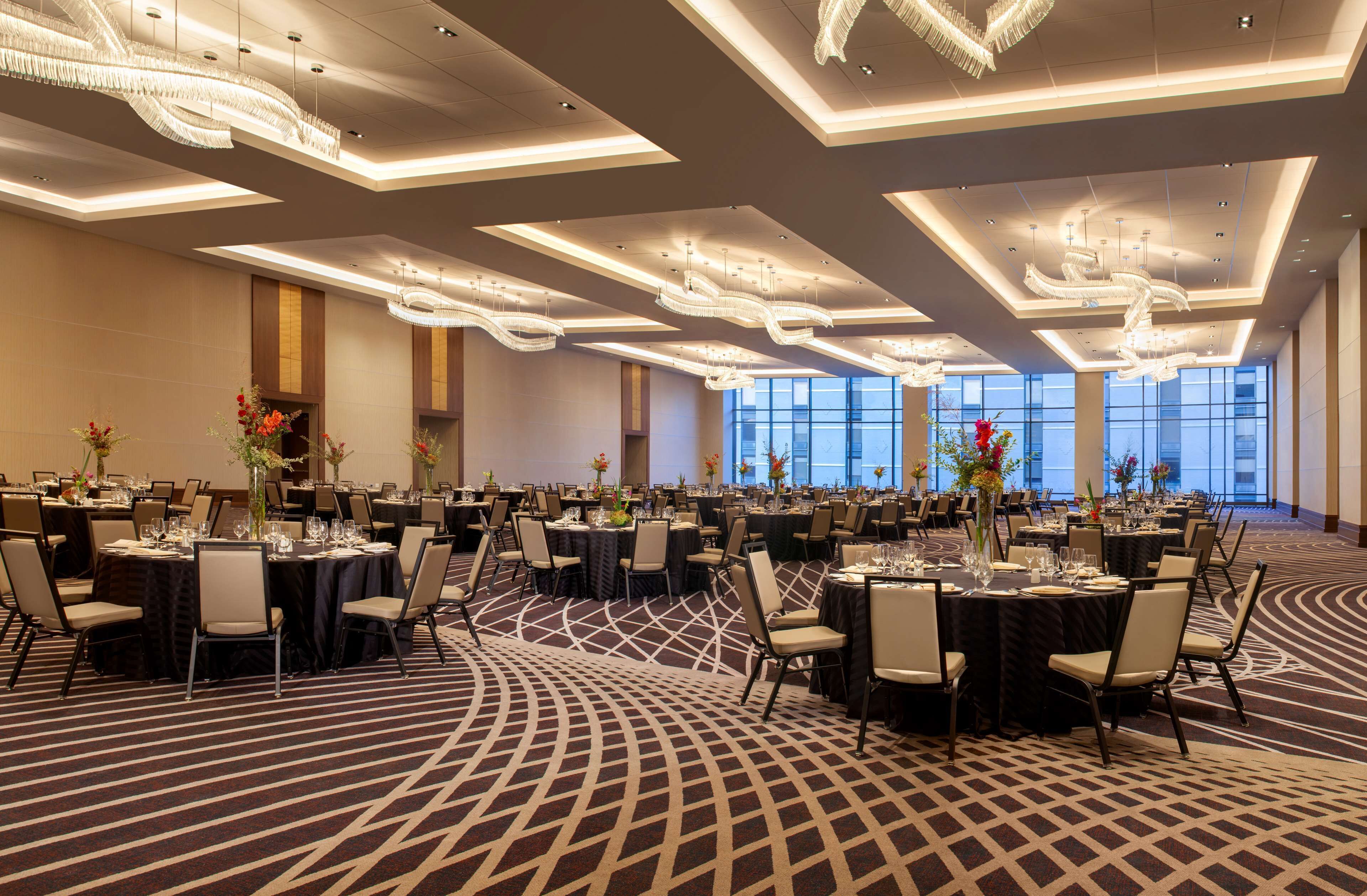Un amplio salón de banquetes con múltiples mesas redondas dispuestas para comer, con una alfombra de estampado geométrico y modernas lámparas de araña.