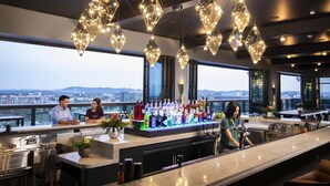 Un bar moderne avec un barman, un comptoir bien garni de bouteilles et de grandes fenêtres donnant sur la ville.