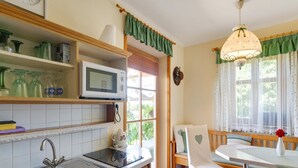 Una cocina con microondas, lavabo y armarios de madera. Hay una ventana con cortinas y una silla cerca.