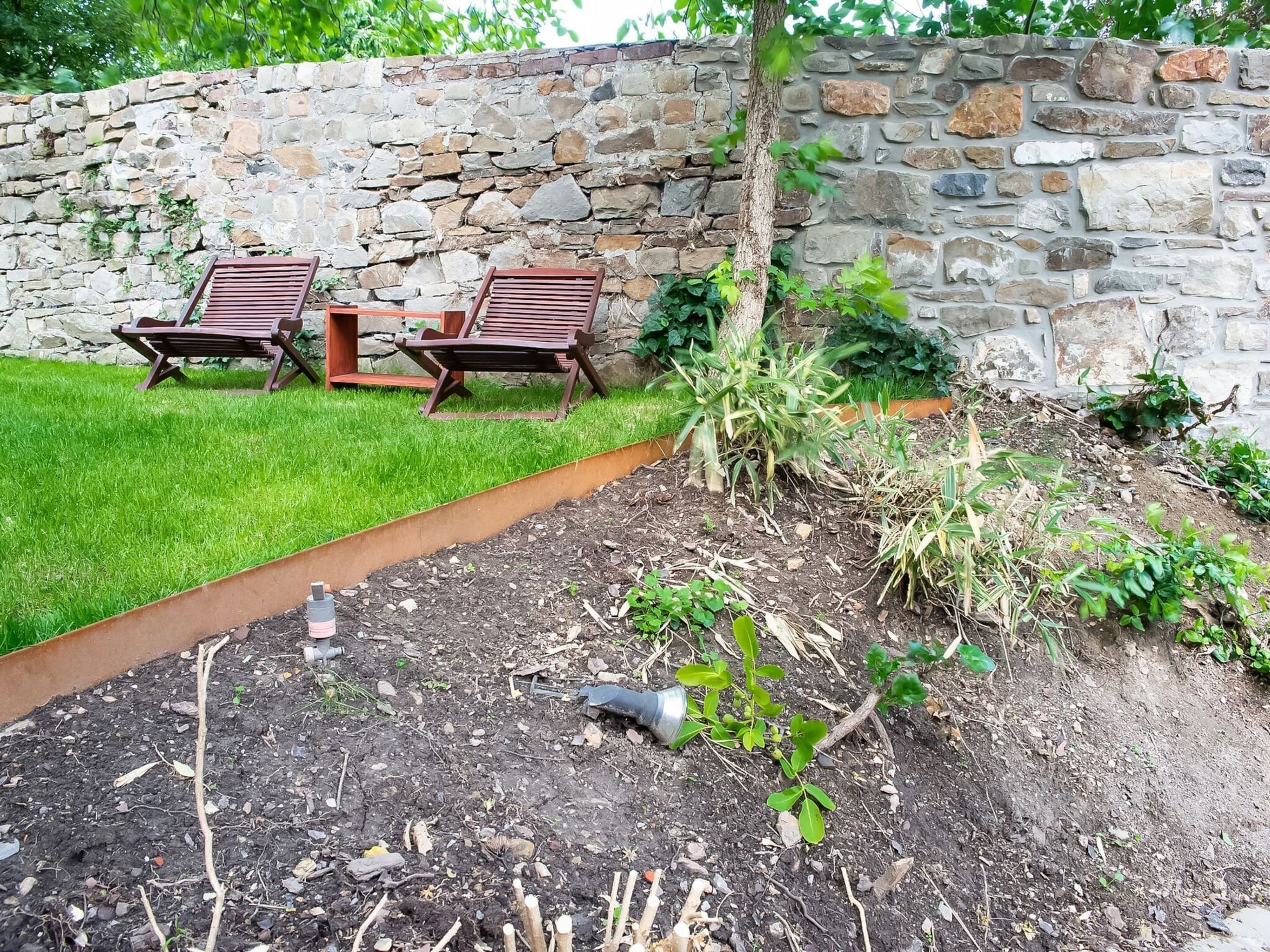 A stone wall, two wooden lounge chairs, a grassy lawn, and a garden bed with various plants.