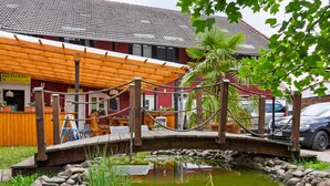 A wooden bridge over a pond with rocks and plants, a restaurant with outdoor seating, and a building with a tiled roof. - Ferienhaus in Meisdorf (Falkenstein)