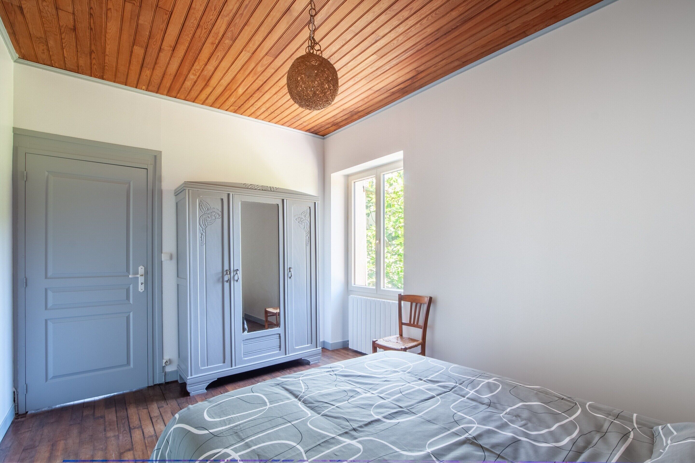 Un dormitorio con techo de madera, un armario grande, una cama con colcha estampada y una ventana con vistas al verdor.