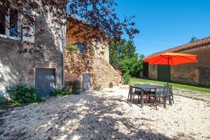 En rustik bygning i sten med en grusbelagt terrasse, udendørs møbler og en rød parasol.