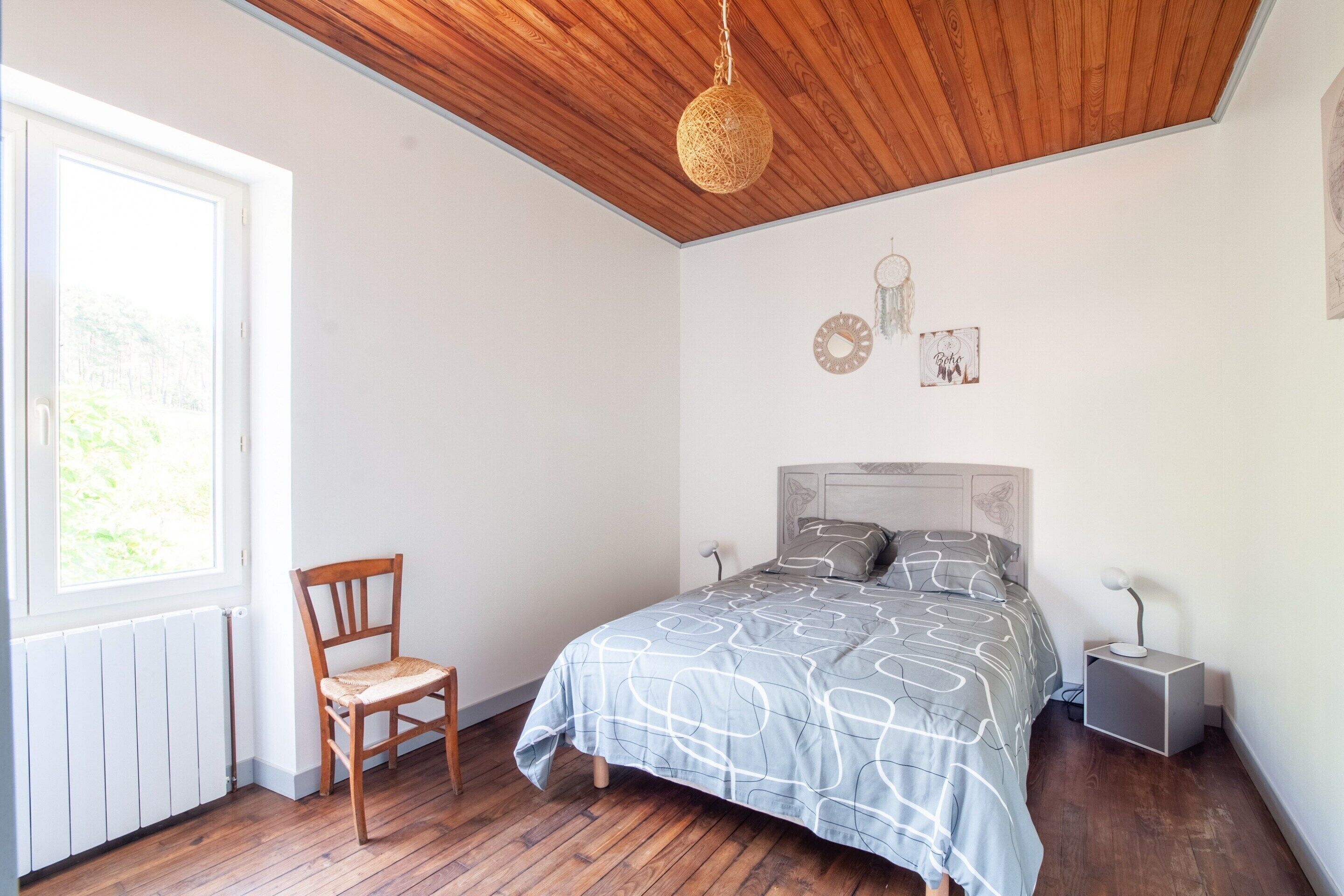 Ein Schlafzimmer mit Holzboden, einem Bett mit gemustertem Überwurf, einem Sessel, einem Nachttisch mit Lampe und einem Fenster mit Blick ins Grüne.