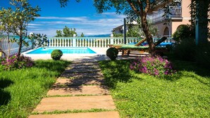 Area piscina con vista aperta sul mare, immersa nel verde e con un vialetto che conduce a un edificio con un balcone.