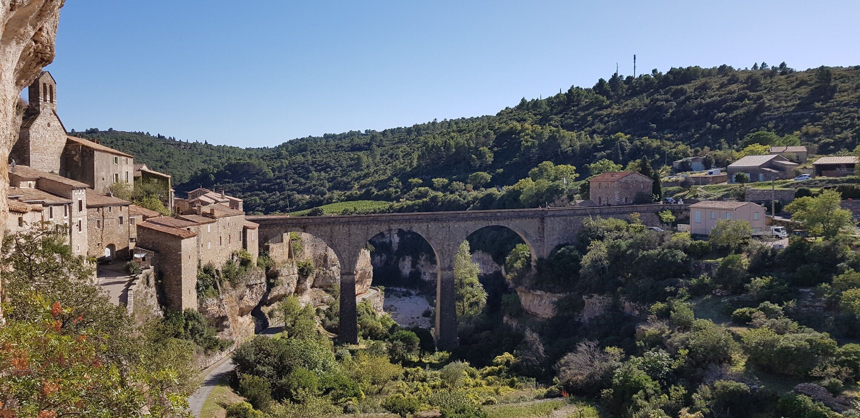 A stone bridge with arches, a stone building with a bell tower, and a hillside with trees.