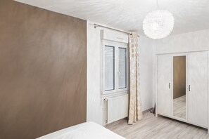 A bedroom with a bed, a wardrobe, a window with curtains, and a hanging light fixture. - Tranquil Garden Retreat by the Sea (Englesqueville-la-Percee)