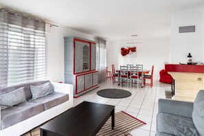 A modern living room with a sofa, a coffee table, a dining area with a table and chairs, and a kitchen area with a red cabinet. - Tranquil Garden Retreat by the Sea (Englesqueville-la-Percee)