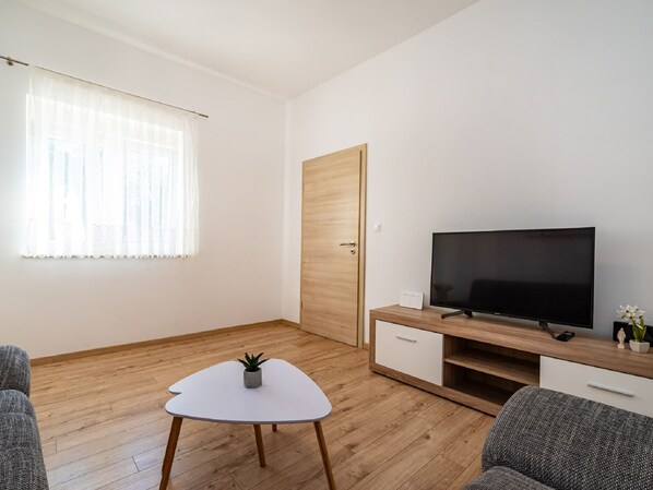 A modern living room with a flat-screen TV on a wooden stand, a small white table with a plant, and a grey sofa.