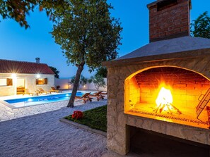 A stone outdoor fireplace with a fire, a swimming pool, and lounge chairs.
