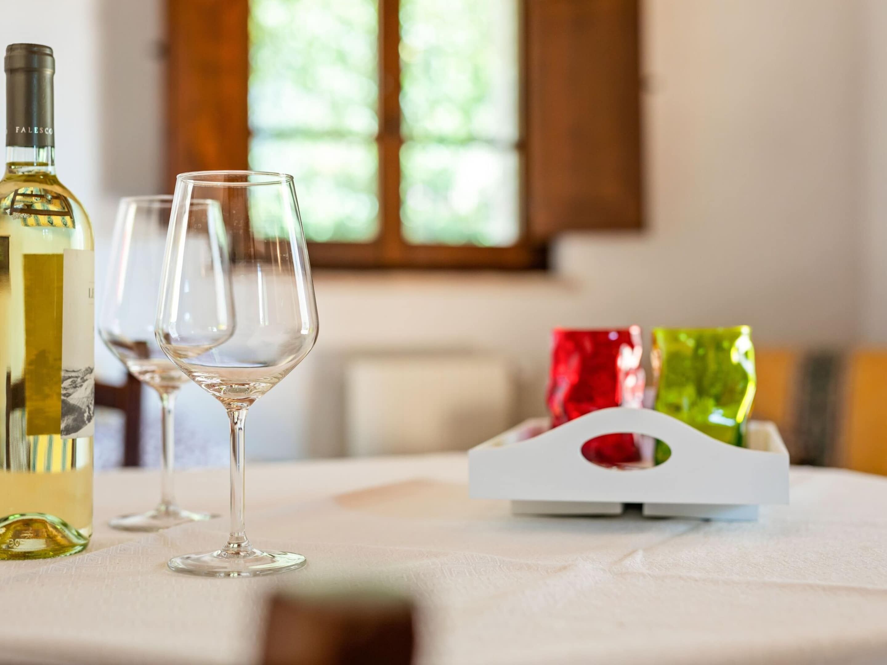 Una mesa con una botella de vino blanco, dos copas de vino vacías y una caja blanca con objetos rojos y verdes.