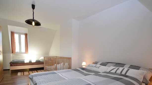 A bedroom with a large bed, a wooden crib, a window with curtains, and a ceiling light. - Comfort in Central Ballenstedt (Ballenstedt)
