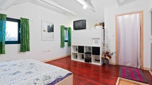 Une pièce avec un lit, un téléviseur posé sur un meuble blanc, un plancher en bois et une porte avec un rideau.