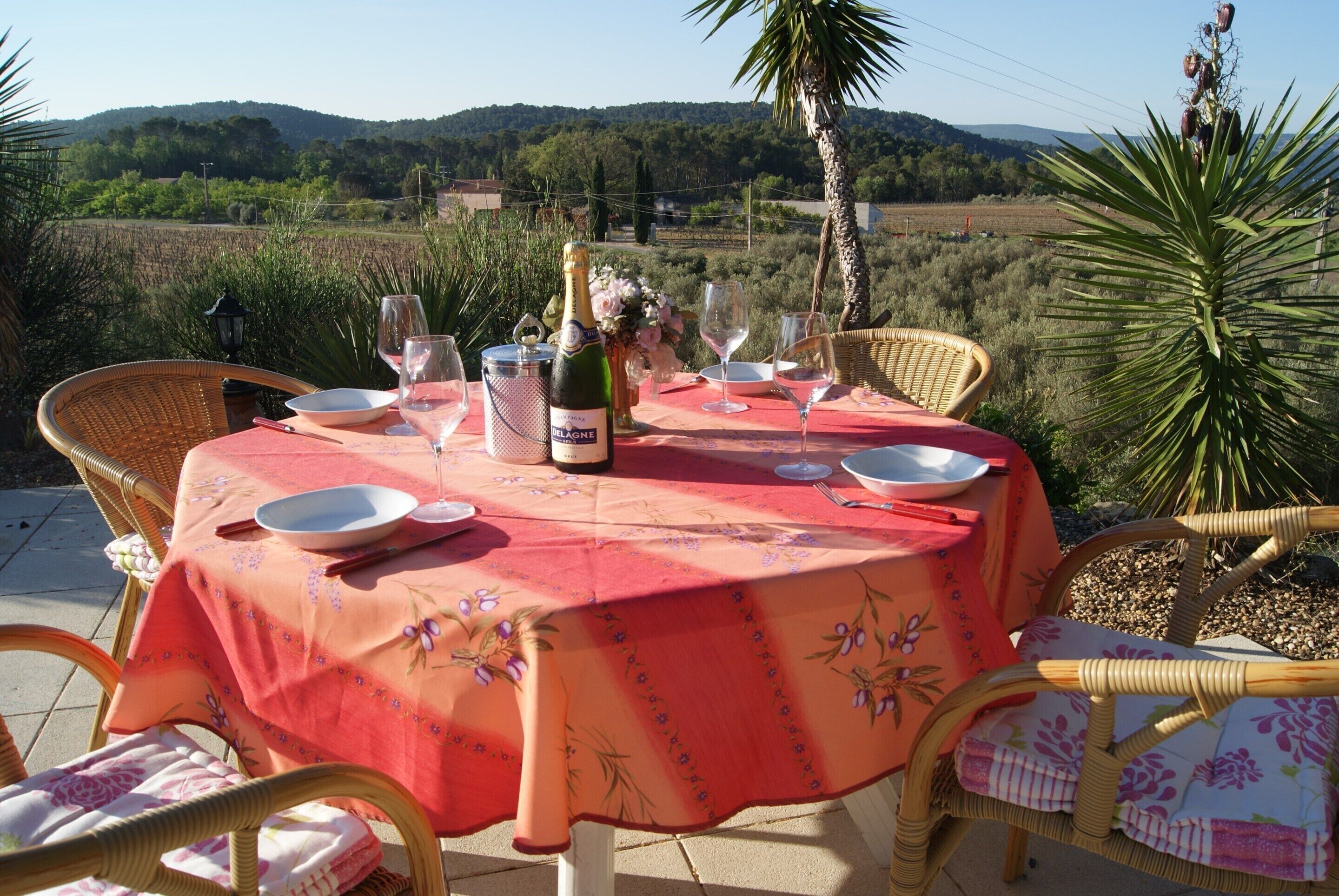 Une table de jardin, dressée pour un repas en plein air, avec une bouteille de champagne, des verres et des assiettes, entourée de chaises en osier.