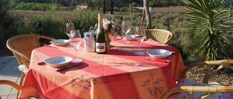 Une table de jardin, dressée pour un repas en plein air, avec une bouteille de champagne, des verres et des assiettes, entourée de chaises en osier.