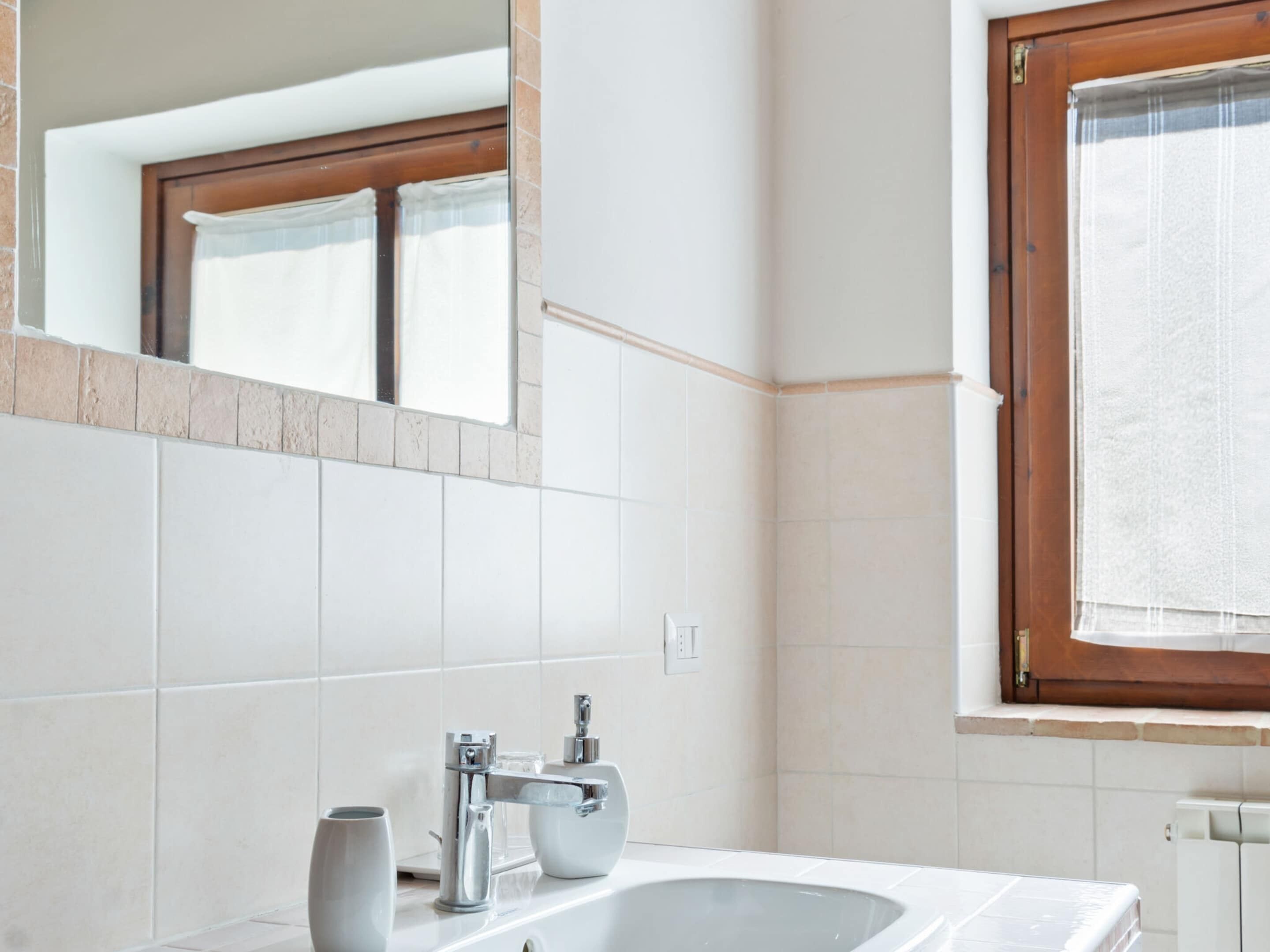 Ein modernes Badezimmer mit weißen Fliesen, einem großen Spiegel, einem Fenster mit Vorhängen und einer Badewanne.
