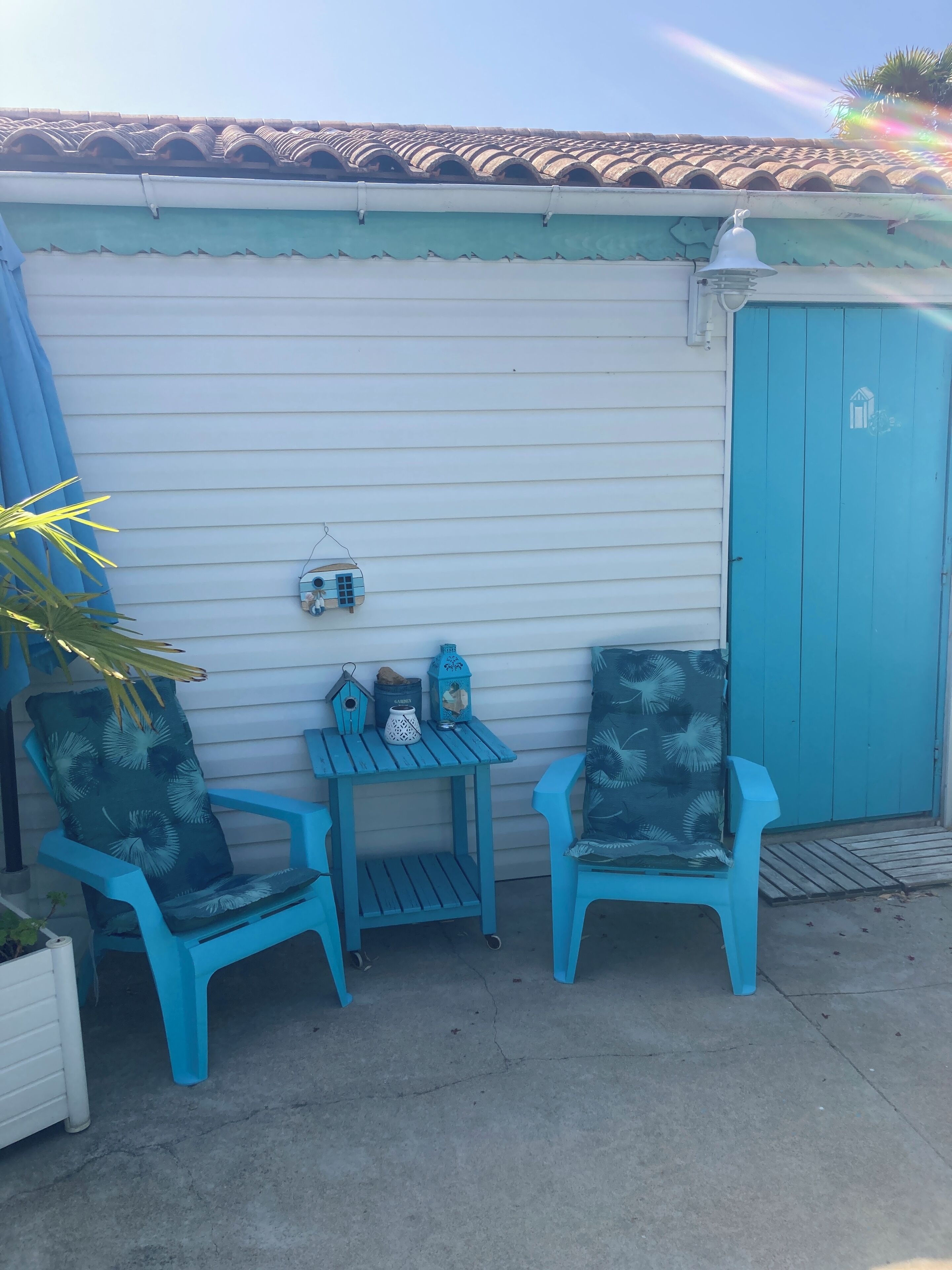 Un espace patio avec deux chaises bleues et une petite table, une porte bleue et un mur blanc surmonté d’un toit en tuiles.