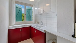 Una cocina con encimeras blancas, armarios rojos, una ventana con persianas verdes y una lámpara de techo blanca.