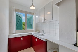 Une cuisine avec des plans de travail blancs, des placards rouges, une fenêtre avec des volets verts et un luminaire blanc au plafond.