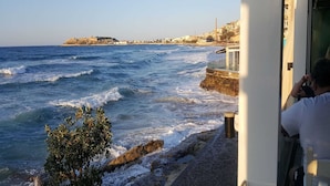 A coastal view with waves crashing onto the shore, a rocky outcrop, and a distant building complex. - Splendid Holiday Home With Swimming Pool (Rethymno)