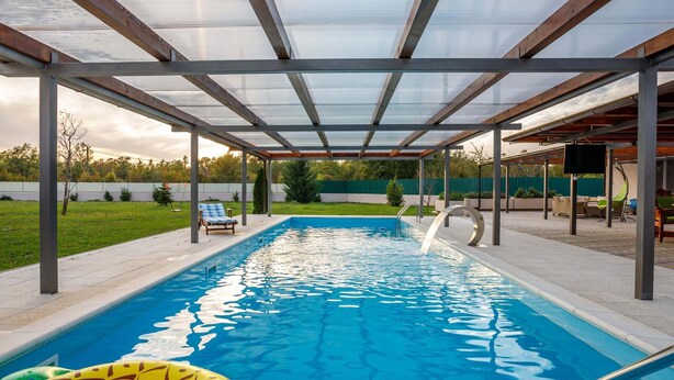 Una piscina cubierta al aire libre con techo de cristal, rodeada por un patio con mobiliario y césped.