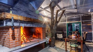 Una chimenea de ladrillo con fuego, una mesa de comedor con sillas y un gran árbol en un ambiente cálido y acogedor.