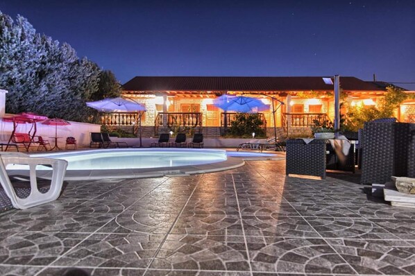 A pool area with lounge chairs and umbrellas, a stone-tiled floor, and a building with a balcony in the background.