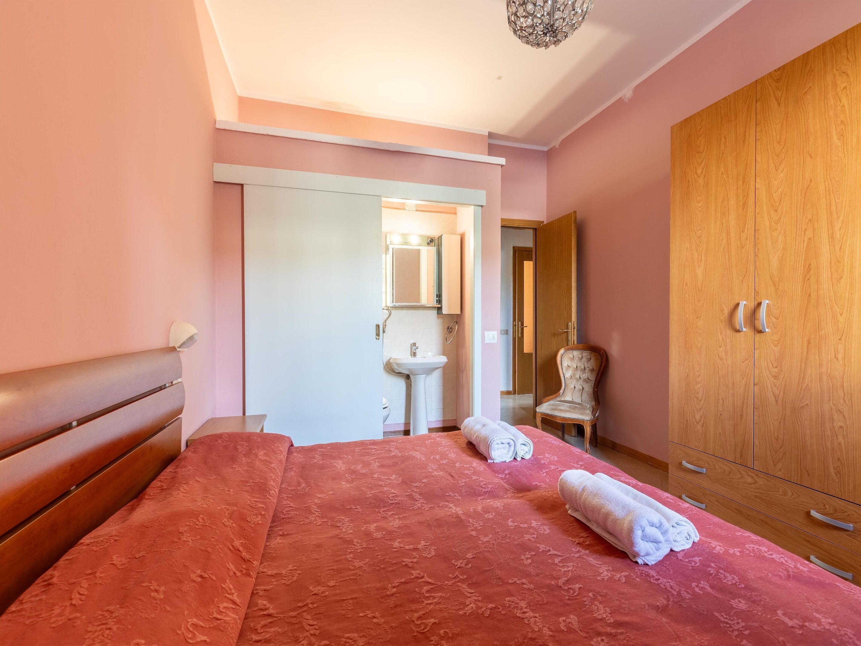 Un dormitorio con cama, un armario de madera, una silla y un baño visible a través de una puerta abierta.