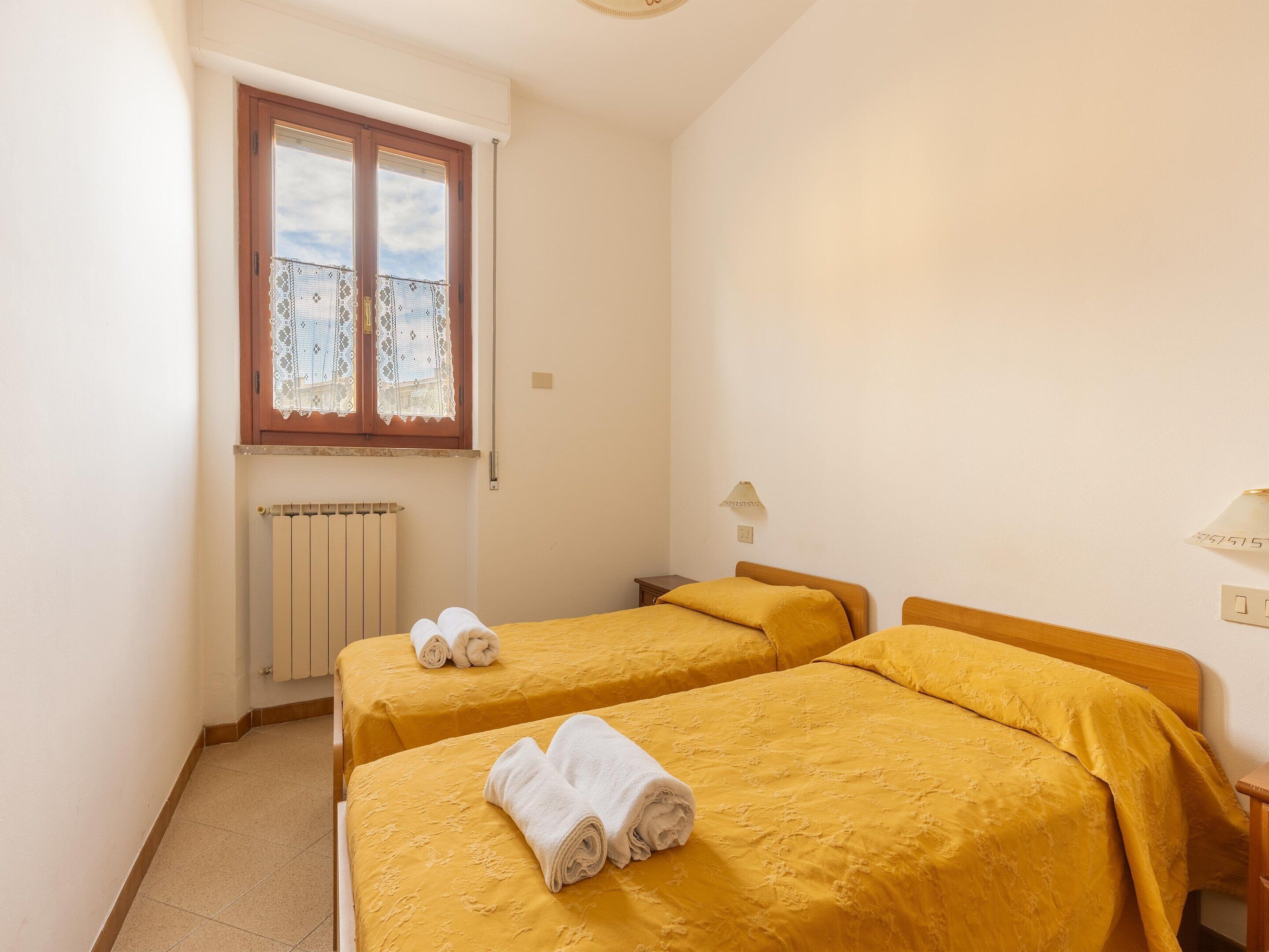 A small, clean room with two single beds, a window with lace curtains, a radiator, and a wall-mounted lamp.
