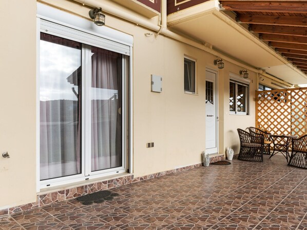 Et overdækket udendørs område med terrasse, skydedør, lille dør og et siddeområde med stole.