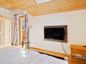 A bedroom with a wooden ceiling, a bed with white bedding, a TV mounted on the wall, and a wooden table.