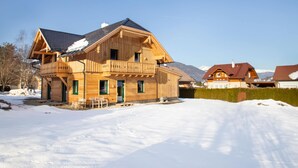A wooden chalet with a balcony, surrounded by snow, with mountains in the background.
