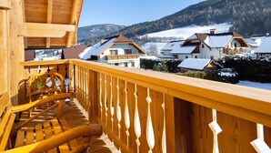 A wooden balcony with a view of snow-covered houses and a forested mountain in the background.