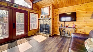 A cozy cabin interior with a stone fireplace, wooden walls, a leather sofa, and a large window overlooking a deck. - Tennessee Chalet (Gatlinburg)