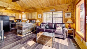 A cozy log cabin interior with a wooden ceiling, a kitchen area, a living room with a leather sofa, and a rustic coffee table. - Tennessee Chalet (Gatlinburg)