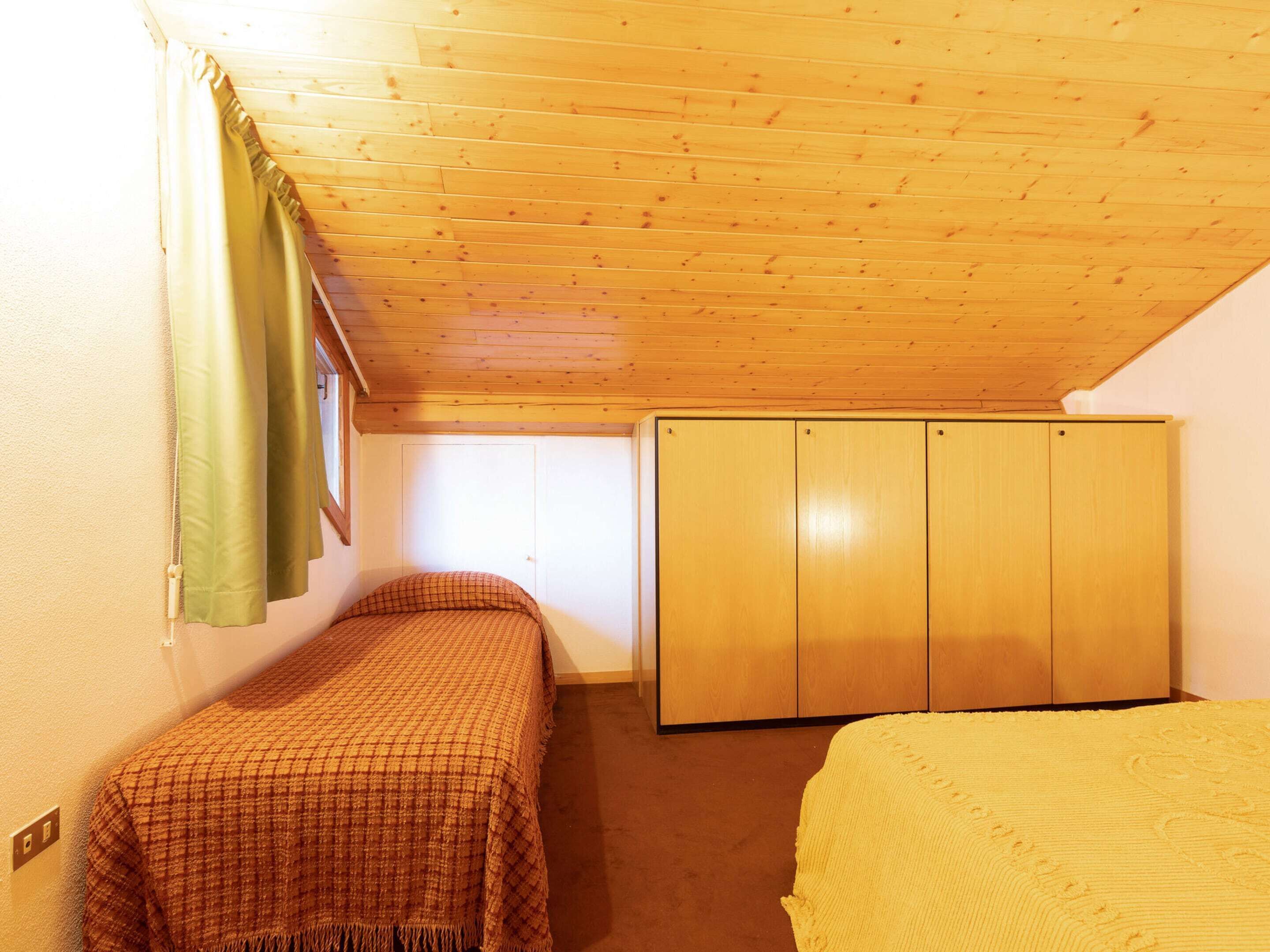 A small, single-room bedroom with a sloped ceiling, a bed, a wardrobe, and a window with curtains.