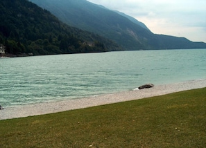 A lake with a pebble beach and grassy area in the foreground, surrounded by mountains. - Castle in Clouds; A Holiday Home (Stenico)