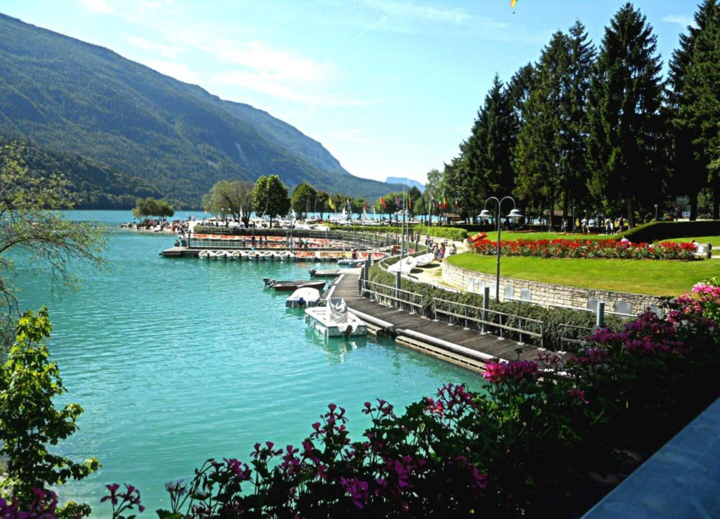 Un parc au bord du lac, avec des bateaux amarrés, une pelouse parsemée de fleurs et une montagne en arrière-plan.