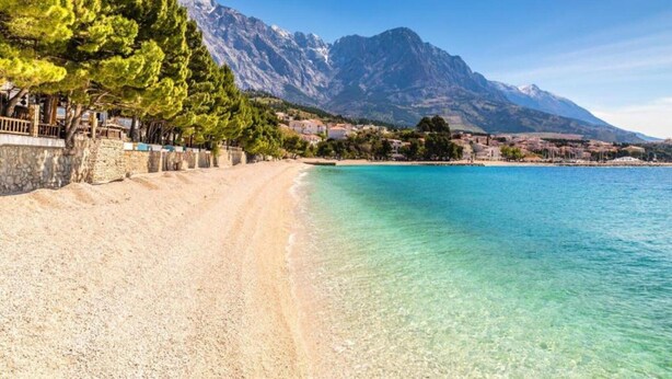 A beach with clear turquoise water, a stone wall, and a mountain in the background.