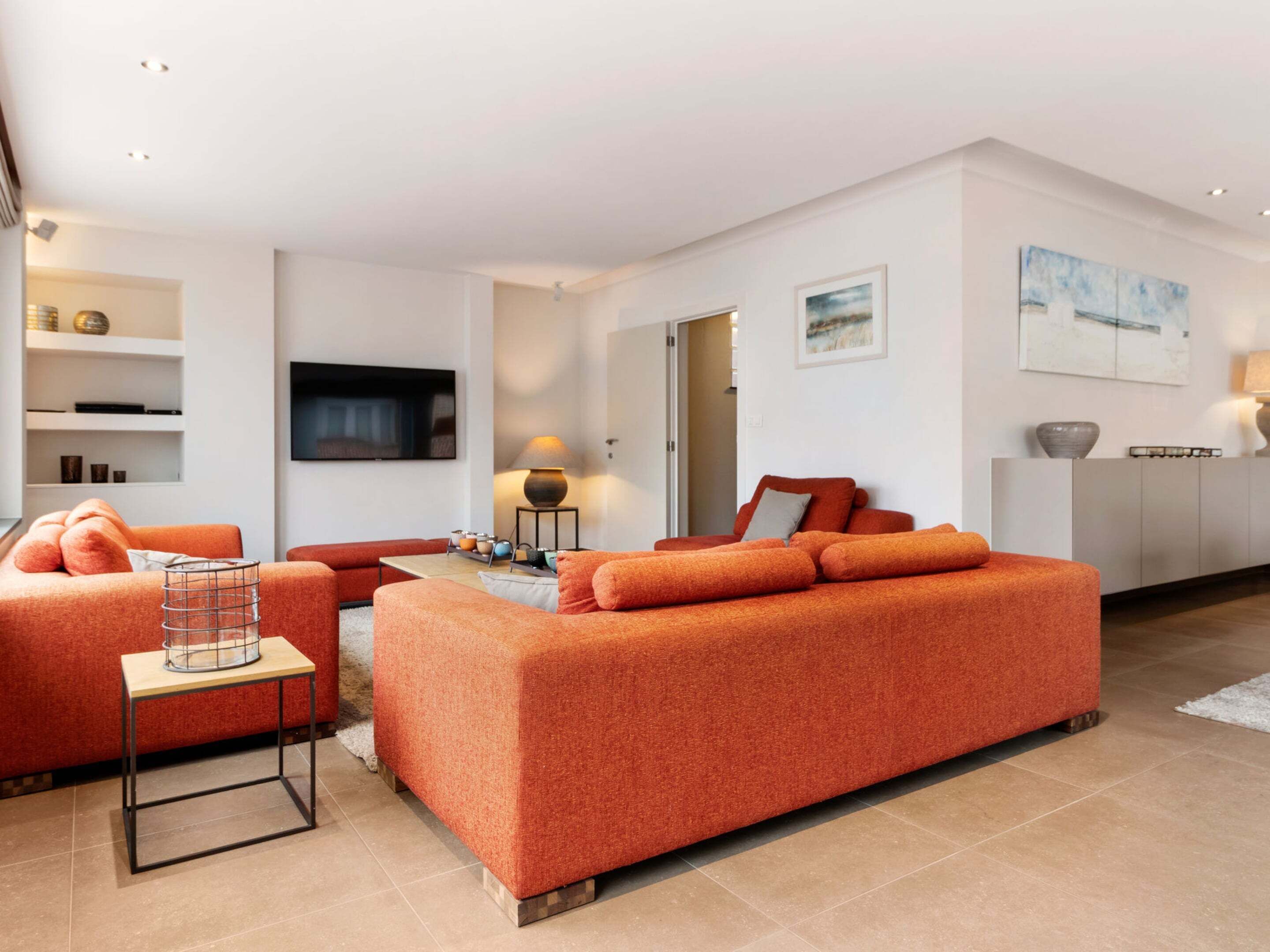 A modern living room with a large orange sofa, a flat-screen TV mounted on the wall, and a minimalist design.