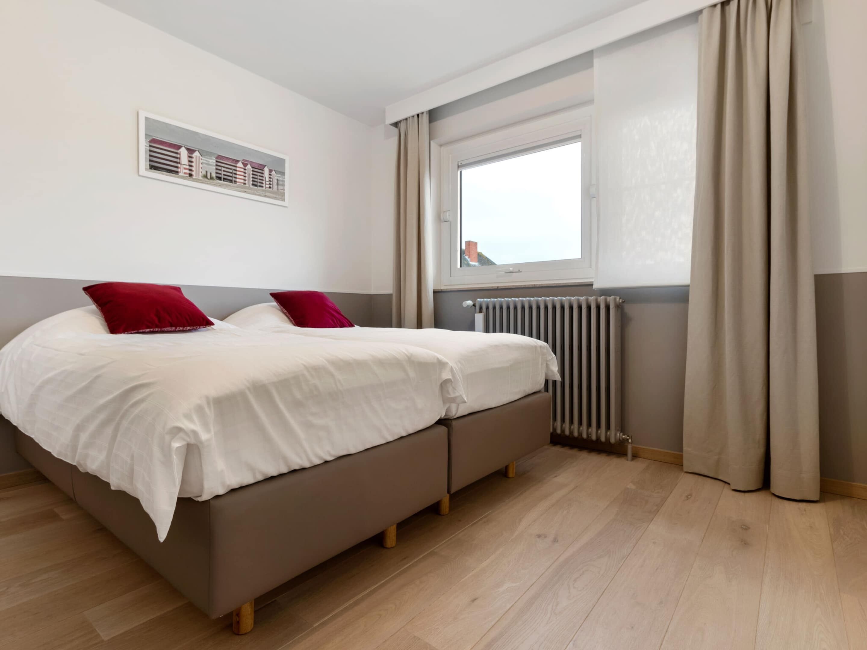 Une chambre moderne avec un lit double, deux oreillers, une fenêtre avec des rideaux, un radiateur et une œuvre d’art accrochée au mur.