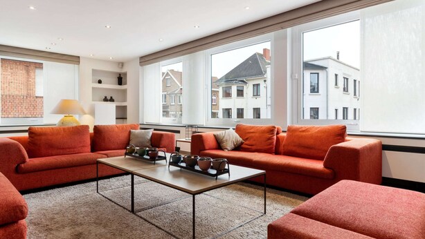 Uma sala de estar moderna com um sofá vermelho, uma mesa de centro e grandes janelas com vista para edifícios.