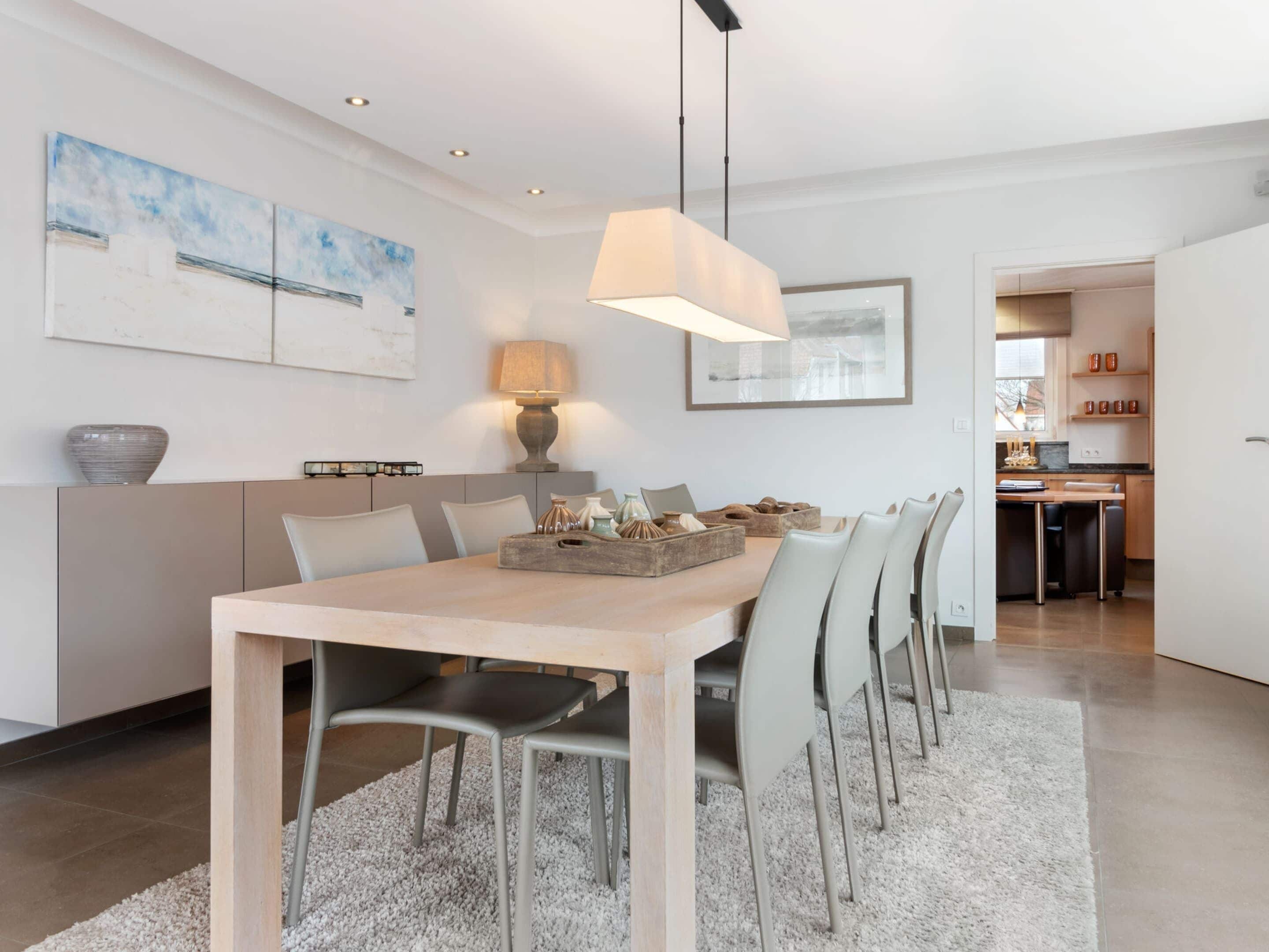 A modern dining area with a light wooden table, grey chairs, a grey sofa, and a white rug. There is a lamp on the table and artwork on the walls.