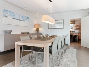 Un espace repas moderne avec une table en bois clair, des chaises grises, un canapé gris et un tapis blanc. Une lampe est posée sur la table et des œuvres d’art ornent les murs.