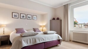 Quarto com cama, duas mesas de cabeceira, guarda-roupa e janela com vista para prédios.